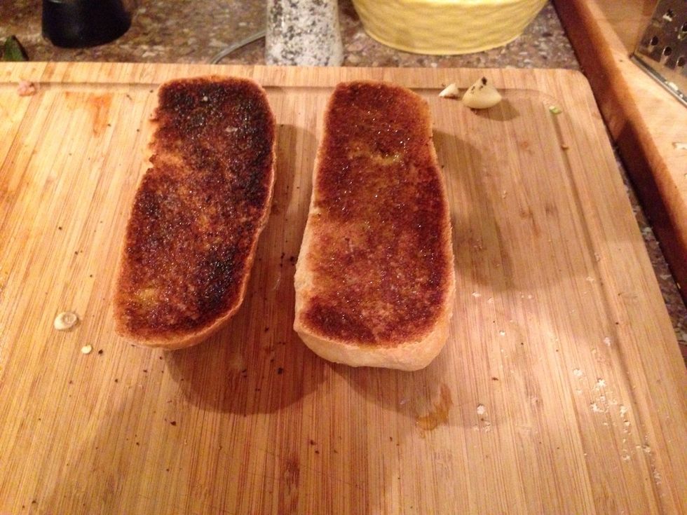 6. Cortamos el pan por la mitad y tostamos cada lado de las rodajas con la parrilla. Cuando est\u00e1n tostados las rodajas, restregamos la otra mitad del ajo por toda la superficie del pan.