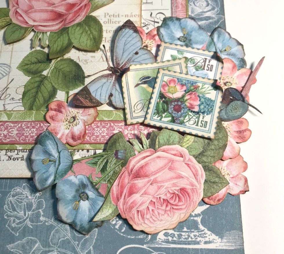 Adhere a pink flower below the 2 pink flowers. Foam square the large pink rose below the stamps. Foam square a blue bunch of flowers to the left of rose. Adhere a pink flower to left of butterfly.