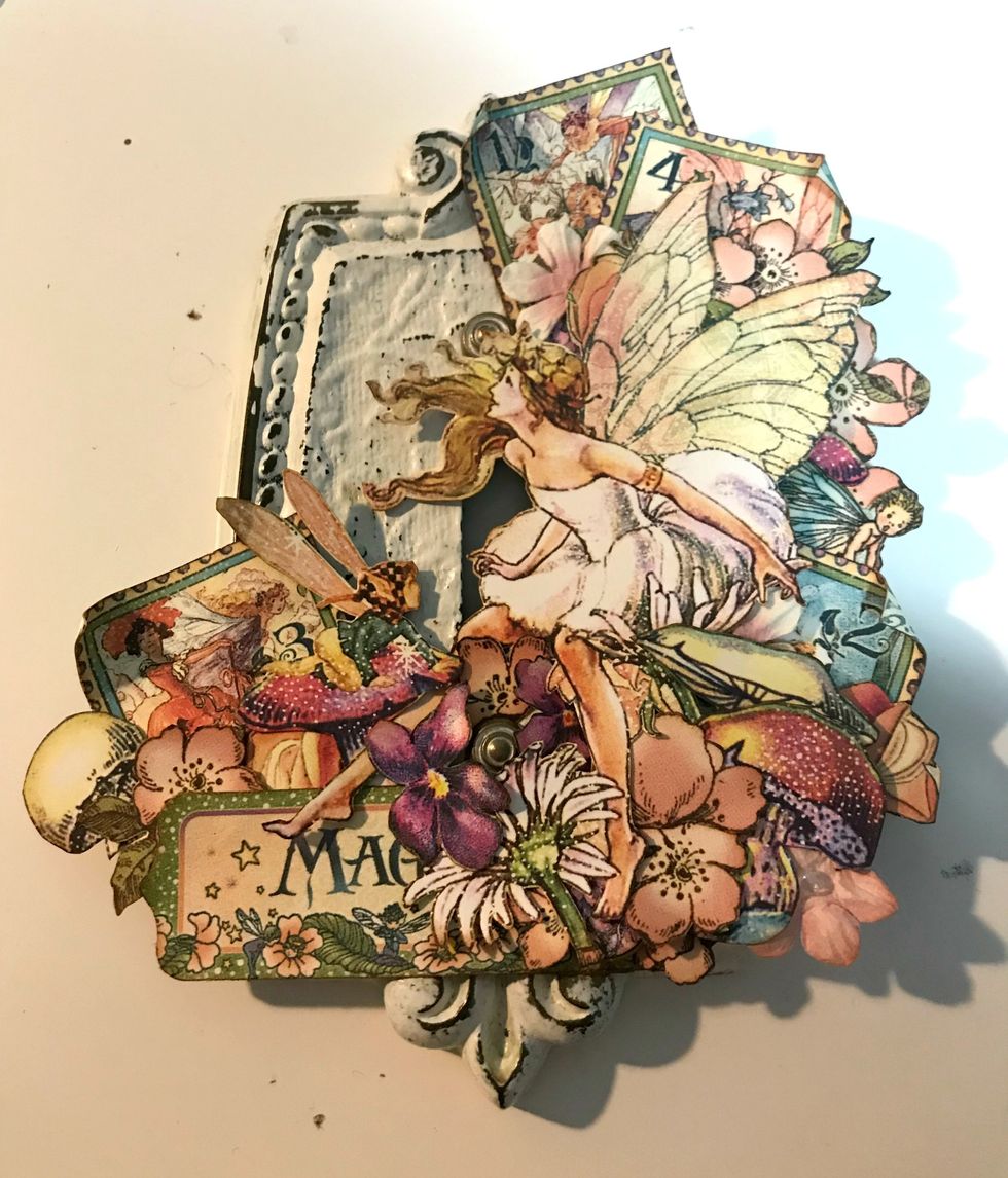 Around the bottom of the fairy, adhere 2 large pink flowers, 2 purple flowers & 1 pink flower. Around the Magic tag, adhere the fairy on toadstool & 1 coral flower. Use foam squares as desired.
