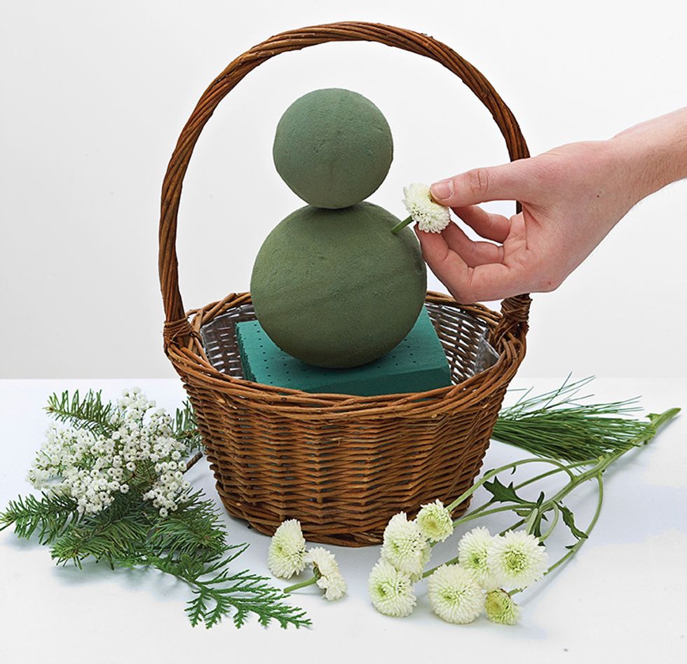Begin by inserting white buttons all around spheres to create the snowman. When Snowman is complete add in Christmas greens around the basket. Insert babies breath around the base of the snowman.