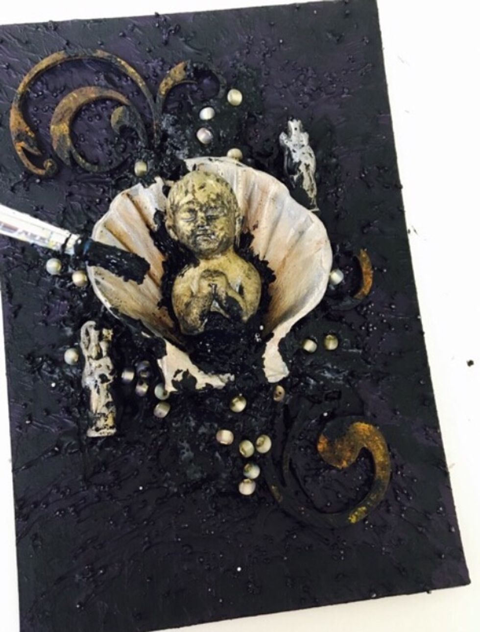 Bring the mixture up into the shell and secure the child figure. Let the texture fan out from under. Add little beaded \u2018gems\u2019 randomly around the shell.