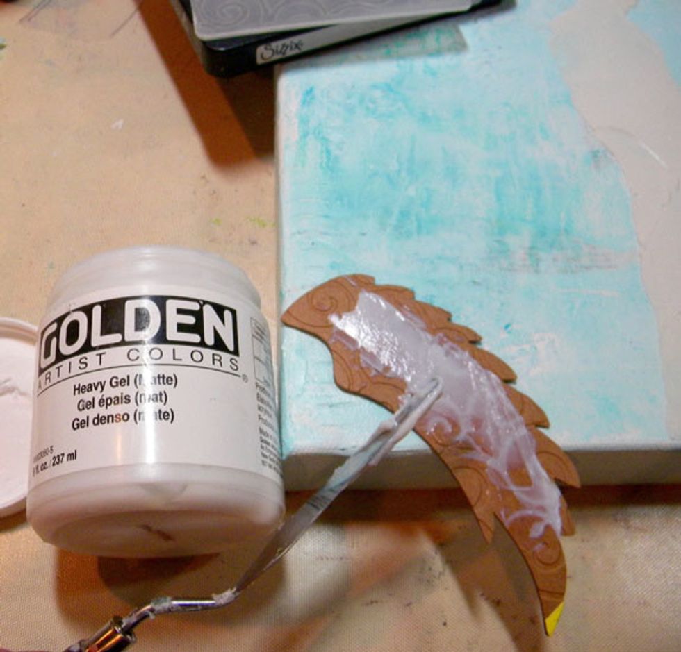 Cut out a wing using a piece of chipboard, the Sizzix Bigz Angel Wing Die, and emboss it with the Cuttlebug D'vine Swirls embossing folder. Glue it down to the canvas using Heavy Gel Medium.