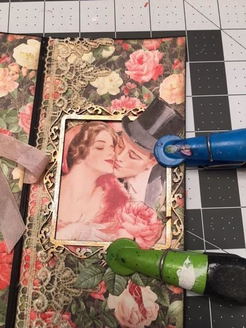 Cut the couple out of the 8x8 Mon Amour paper. Arrange them in and out of a chipboard frame that has been inked with silver and gold.  Glue the image and frame down. Use clips to hold until dry.