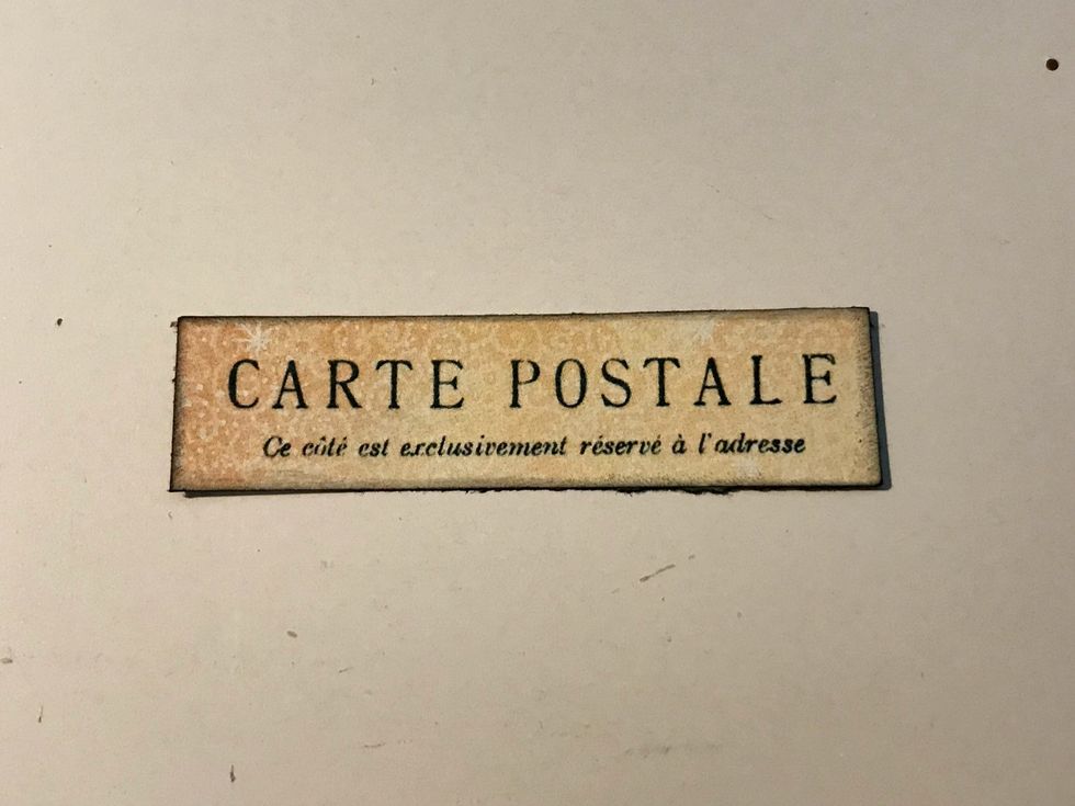 From the backside of another ephemera card, cut out the Carte Postale. The piece of paper will measure 1/2" x 2 1/2". Ink the edges of the paper using Photogenic Ink and a blending tool.