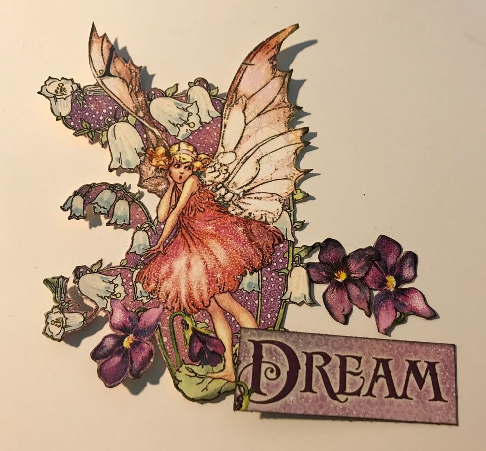 From the card paper, fussy cut the fairy and two purple flowers. From the backside of the ephemera card, cut the dream word out. Ink the edges of the pieces using Photogenic Ink and a blending tool.