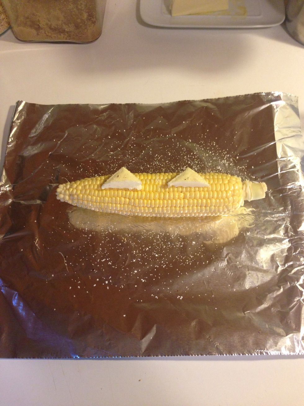 I take a pat of butter, cut it in half, and place it on the corn like so. Then I take some seasoned salt and pour it over the top. Doesn't matter if it goes everywhere, it's all getting wrapped up.