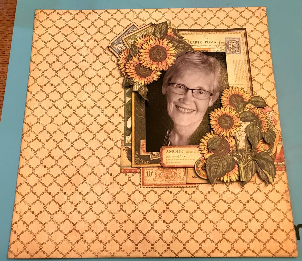 In the bottom right hand corner of the photo, use foam squares and adhesive to adhere the Amour sticker, 3 sunflowers, sunflower bunch, rooster and two sunflower chipboard buttons.