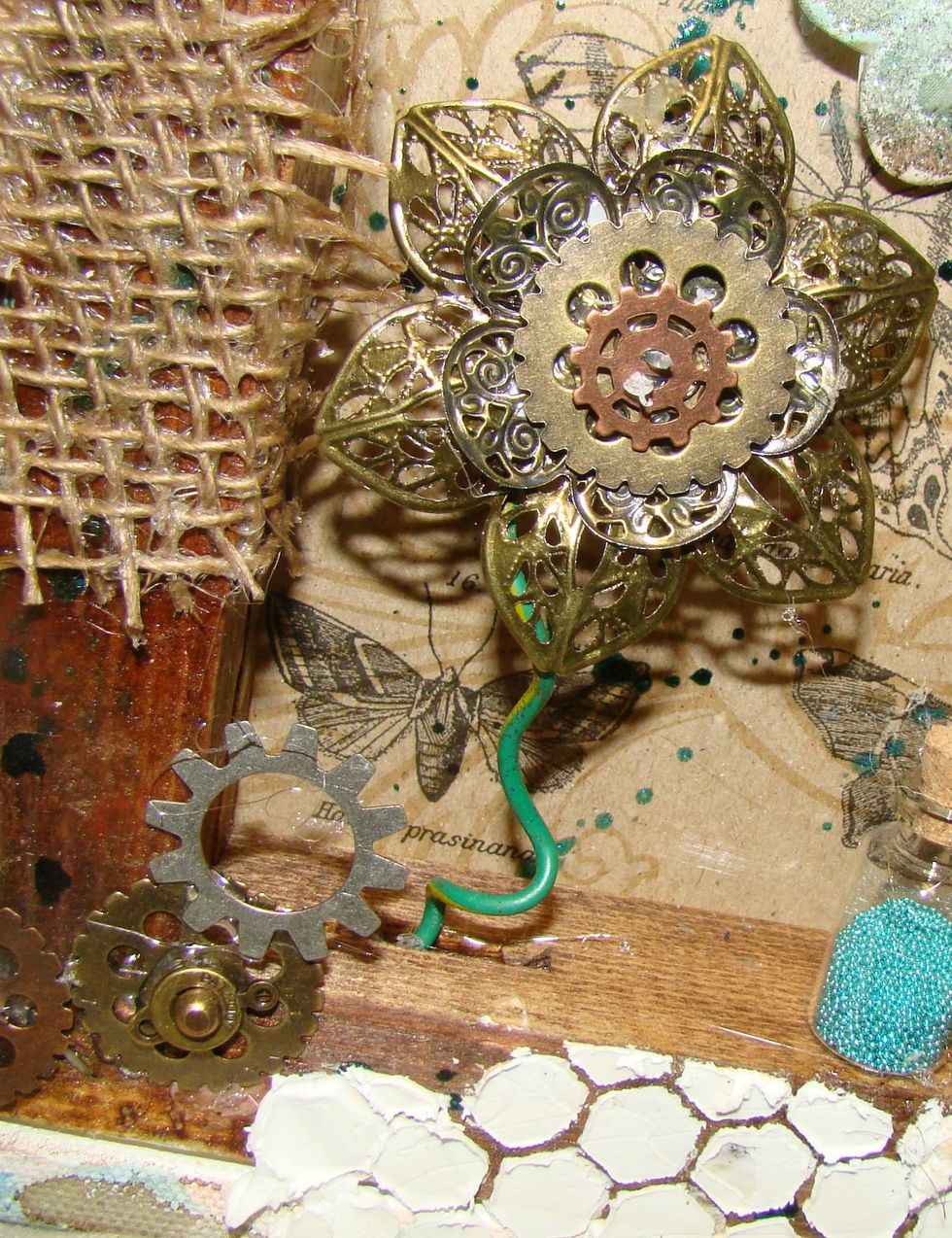 Layer different metal flowers on top of each other and add cogs and gears to create your flower. Glue the stem to the flower and glue to your wood frame portion of the canvas.