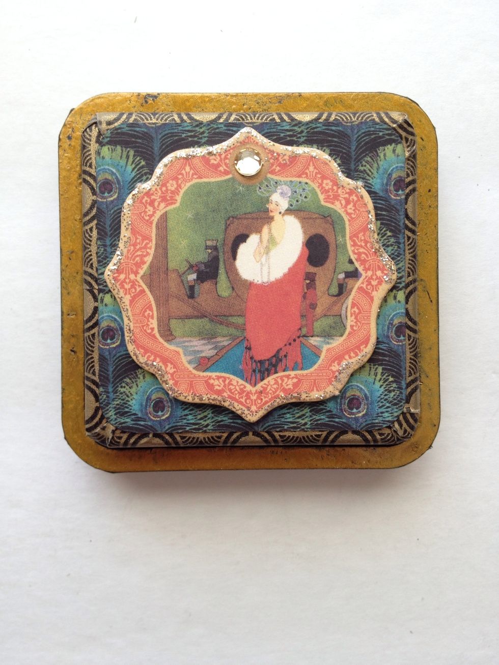 Locate the chipboard piece in the photo and adhere to the top of your lid. Attach a crystal and apply some Stickles around the chipboard.
