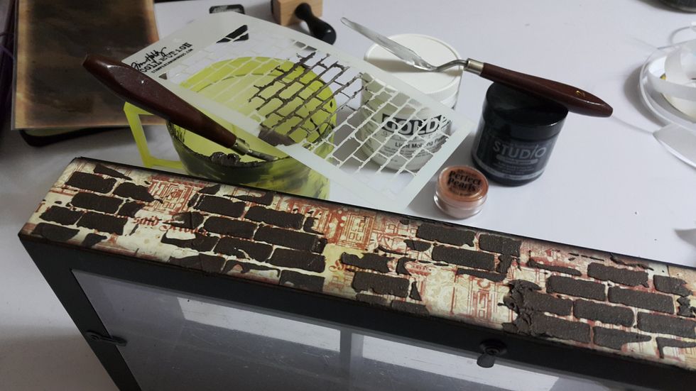 Mix Light Molding Paste, Black Gesso and Perfect Pearls for the bricks color, spread randomly with stencil by the side and on the top of the window shadow box