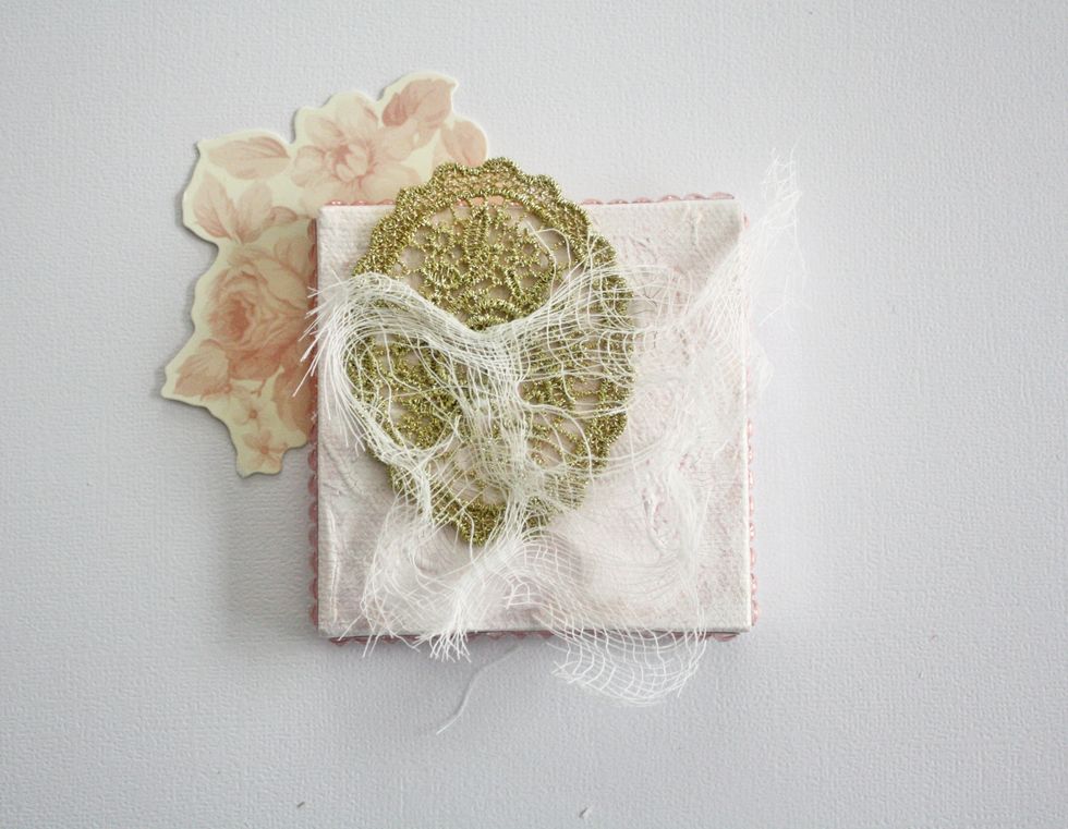 Now to start layering the embellishments. The floral die cut is adhered to the back of the canvas. Cut a piece of the Prima trim and layer cheese cloth on top.