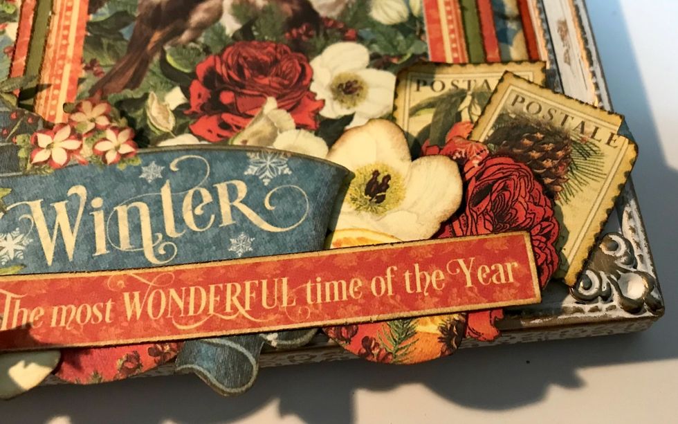 On the right side of the winter banner, adhere 2 red roses, 1 white flower and two stamps. Roll one of the edges down on each corner of the stamps.