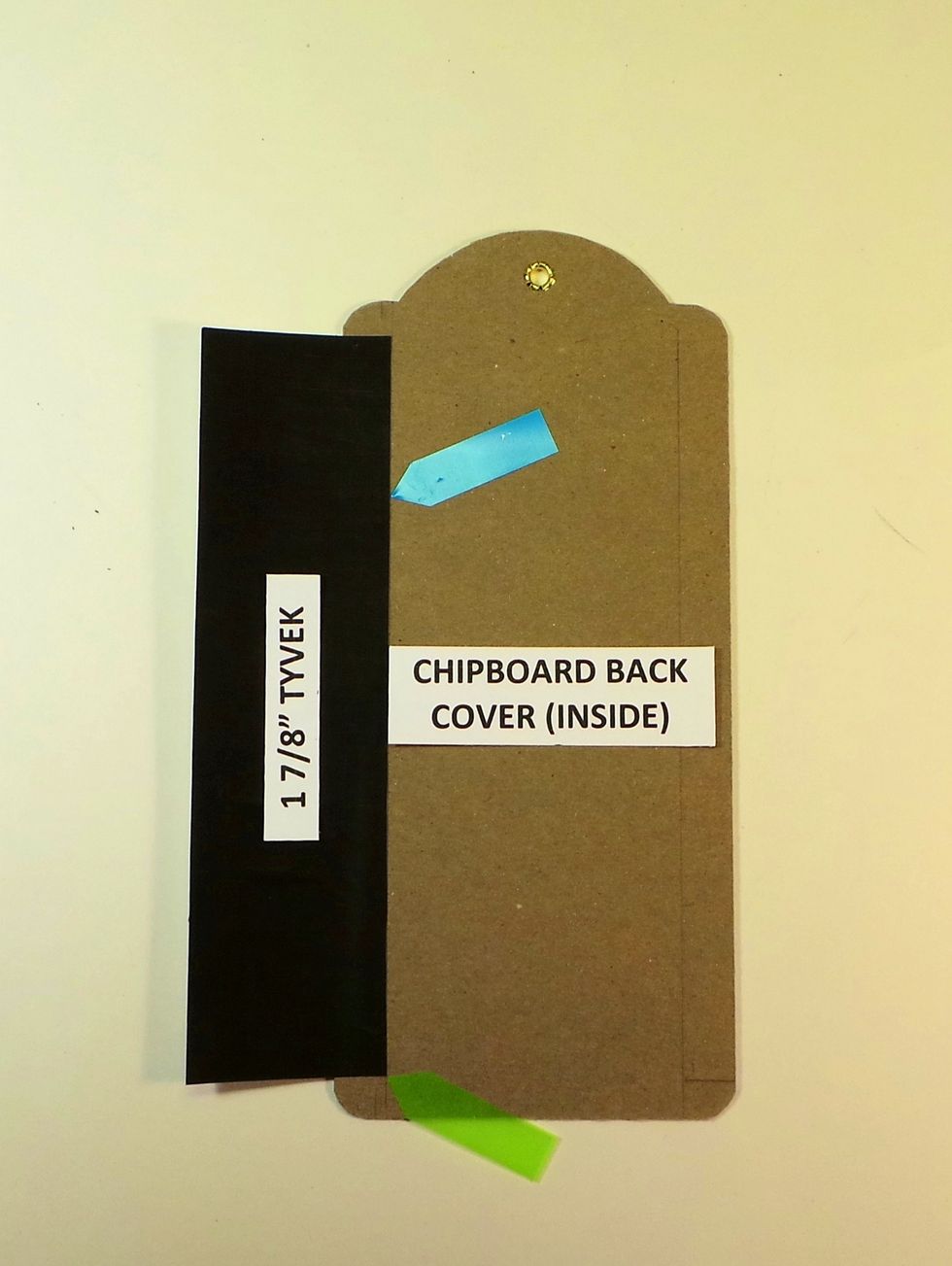 Place a Chipboard tag with inside facing up. Remove 1/2" tape backing from 1 edge of 1 7/8" Tyvek Align bottom corner with 3/8" line (green arrow), right edge with 1/2" line (blue arrow). Burnish