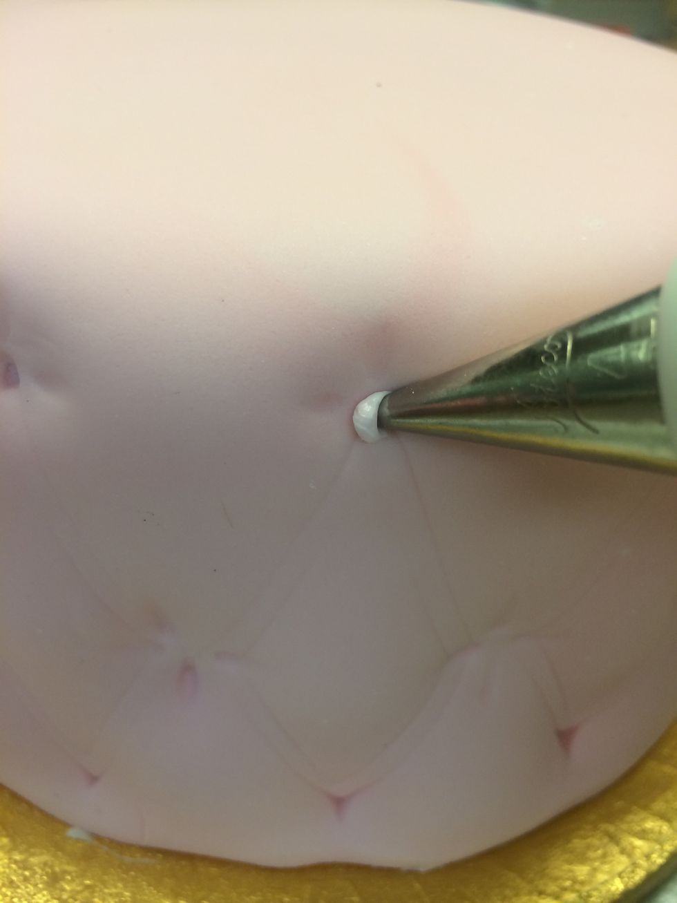 Prepare a pastry bag fitted with a round tip. Put your royal icing in the bag. Pipe a tiny amount into the round small center stitch. Then place a gold bead onto each piped pink dot.