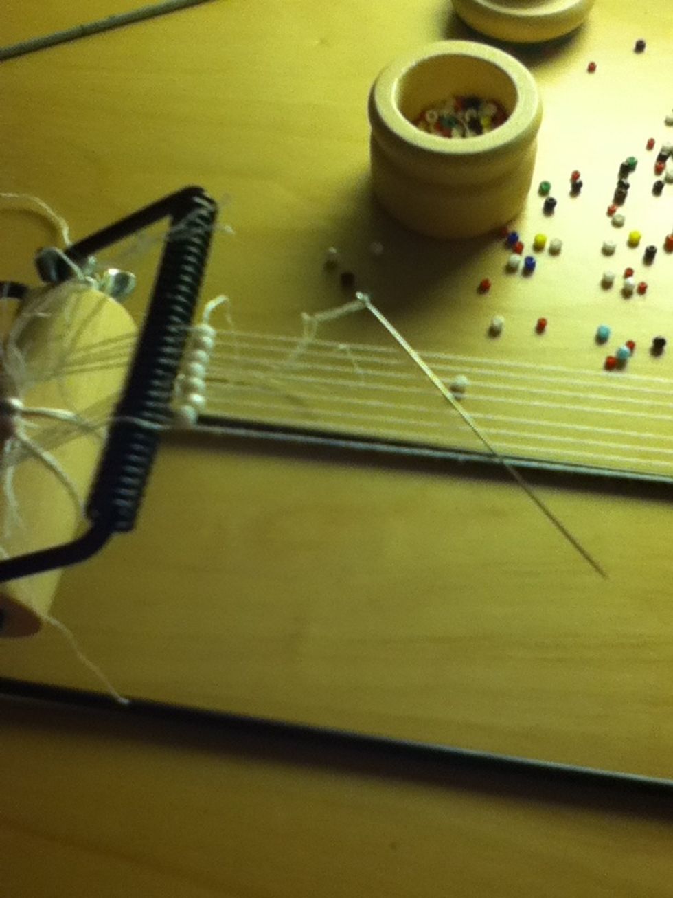Push the beads into the separate holes with your palm and then put the needle and thread under the loom and through the beads, but on TOP of the loom thread. Here is a video.