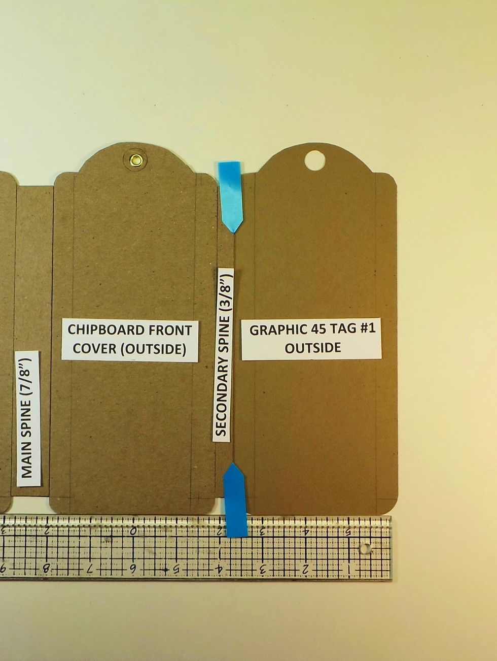 Remove backing from remaining Scor-tape and butt the a Graphic 45 paper tag against the spine (blue arrows). Outside of tag should be facing up. Use a straight edge to keep everything in alignment.