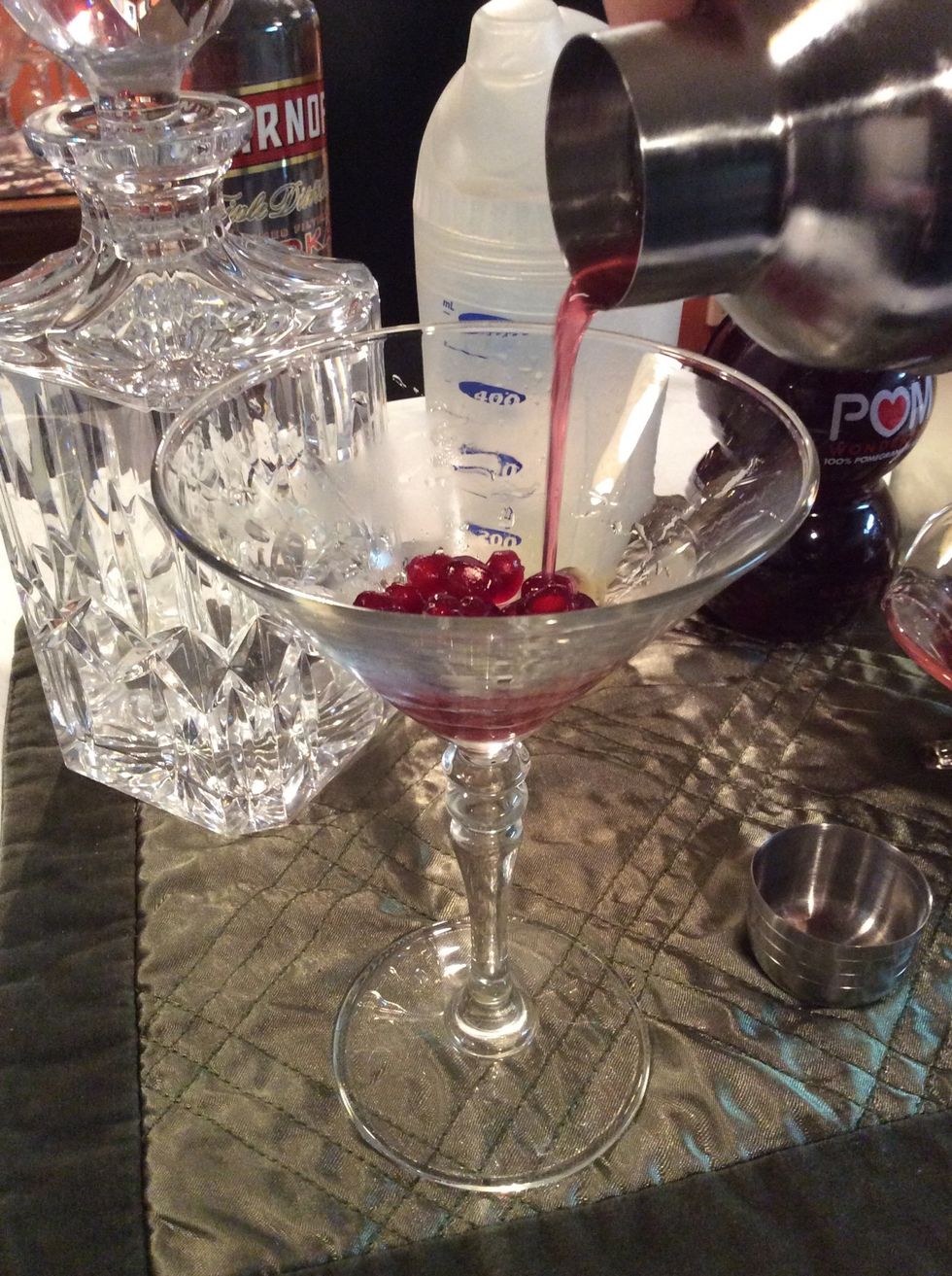 Remove cap and strain over chilled martini glass with awaiting pomegranate seeds