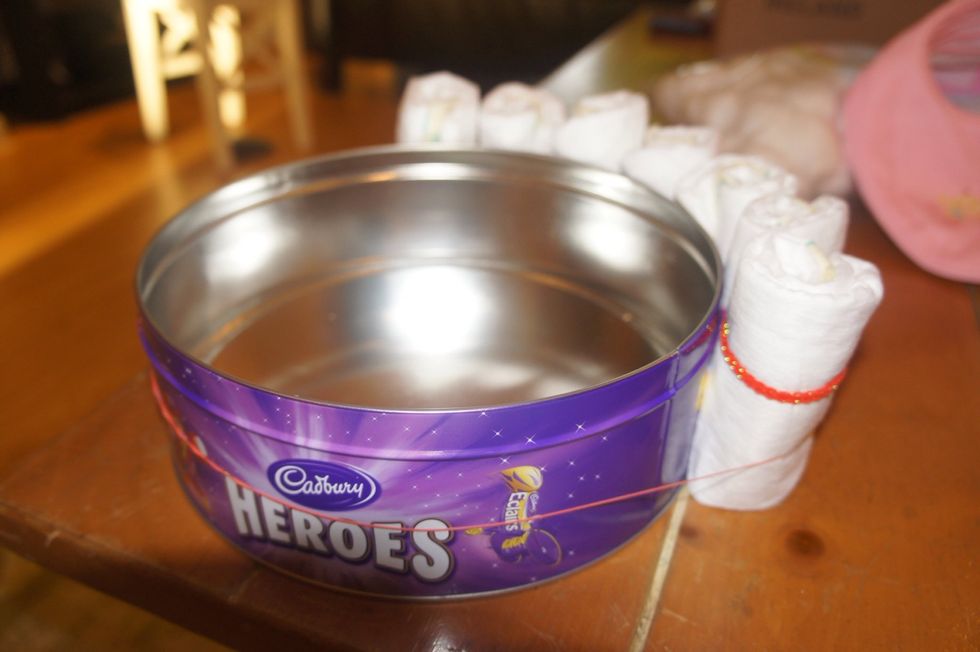 Remove the lid from empty sweet tin. Place a large elastic band around the tin. Make sure there is a little bit of give, but also it is not too loose. Start loading the rolled nappies in the band.