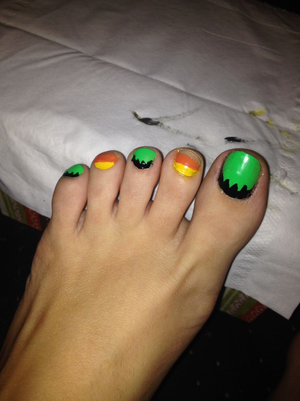 Start with your left pinky toe and paint a black "hair line" on the green nails. Paint a orange strip across the middle of your yellow nails, underneath the yellow. Clean up smudges as you go.
