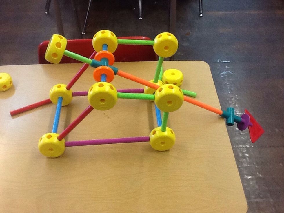 Step 4B:Assemble the launcher by putting one intersection with a blue stick through one side and one orange stick on one side and a green stick with a yellow wheel attached to it.