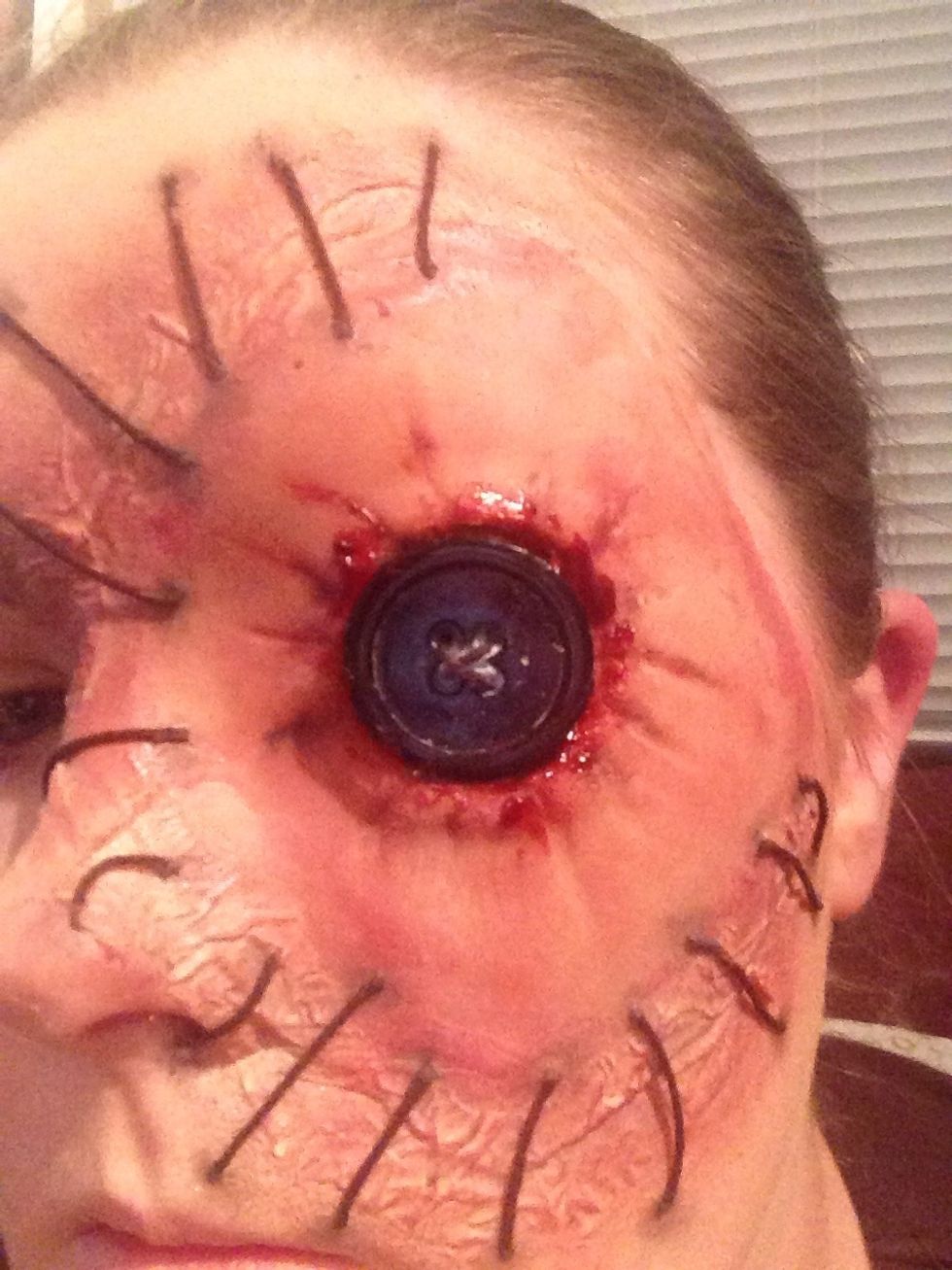 Stick the button on with some liquid latex or spirit gum, and let I dry.  Then apply some thick blood around the edge of the button. Smudge it a bit, look more realistic then.