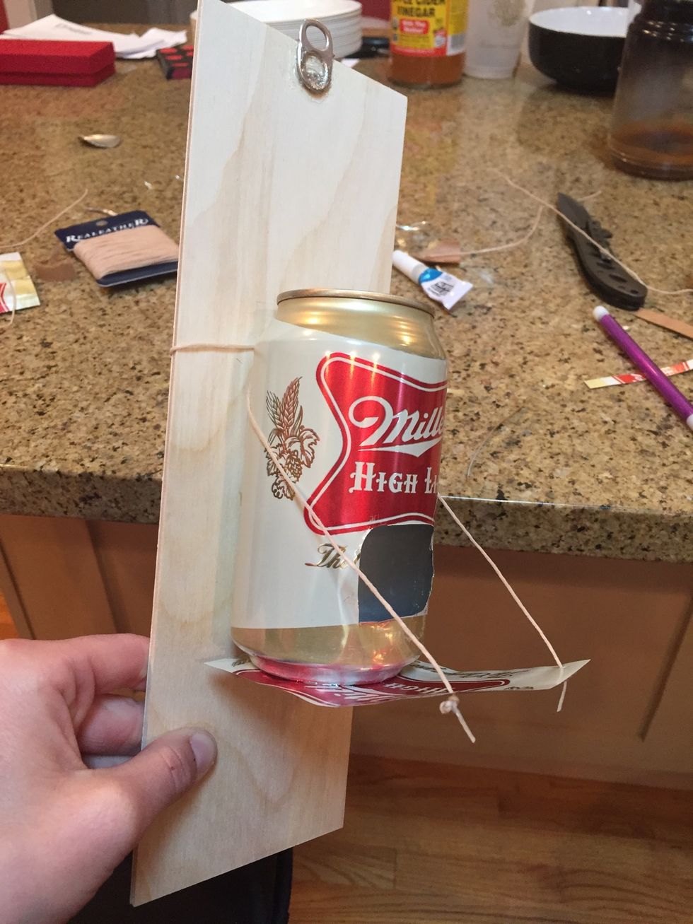 Take both pieces of string and wrap them around the back of the can then wrap the string around the back of the wood