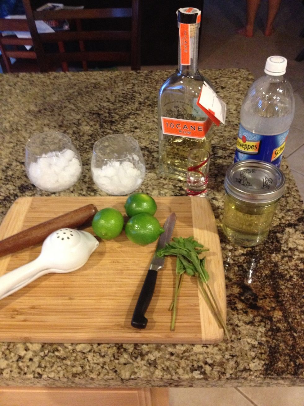 The supplies Rum, simple syrup, mint leaves, limes, club soda, glasses with ice, muddler, citrus juicer and knife