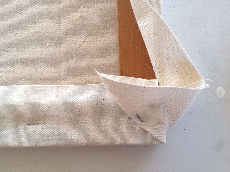 This is similar to wrapping gifts. Pull the corner piece toward the center of the canvas till it makes a fold from the corner of the frame and staple it into position.
