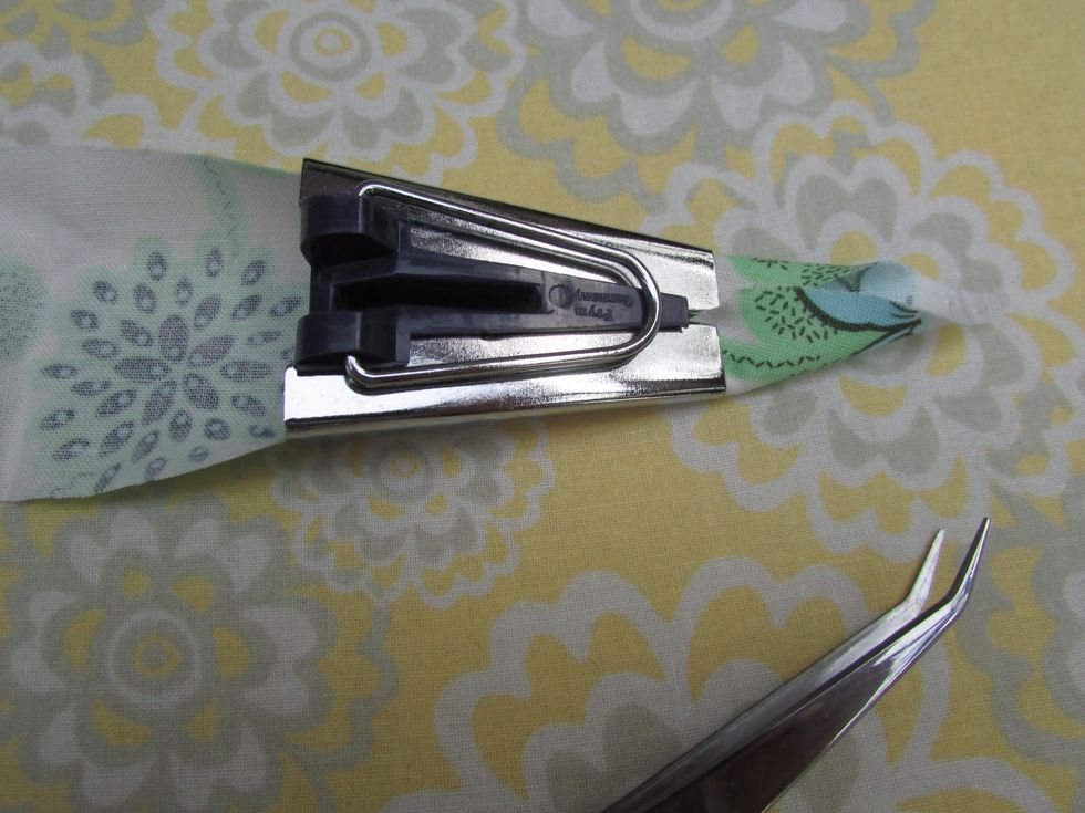 This one inch bias trim tool is what makes the process a whole lot quicker: feed the fabric, pointed end first, wrong side up, through the tool and pull the fabric through to get it started.