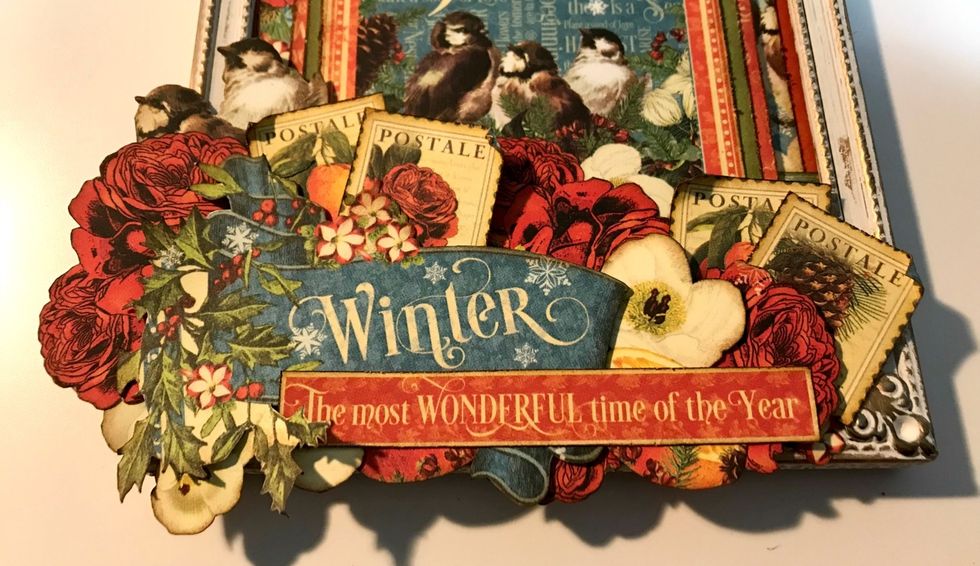 To finish off around the winter banner, adhere two stamps, one bird, one red rose. Roll a corner down on each stamp. Using a few of the small flowers, fill in the bottom of the frame.