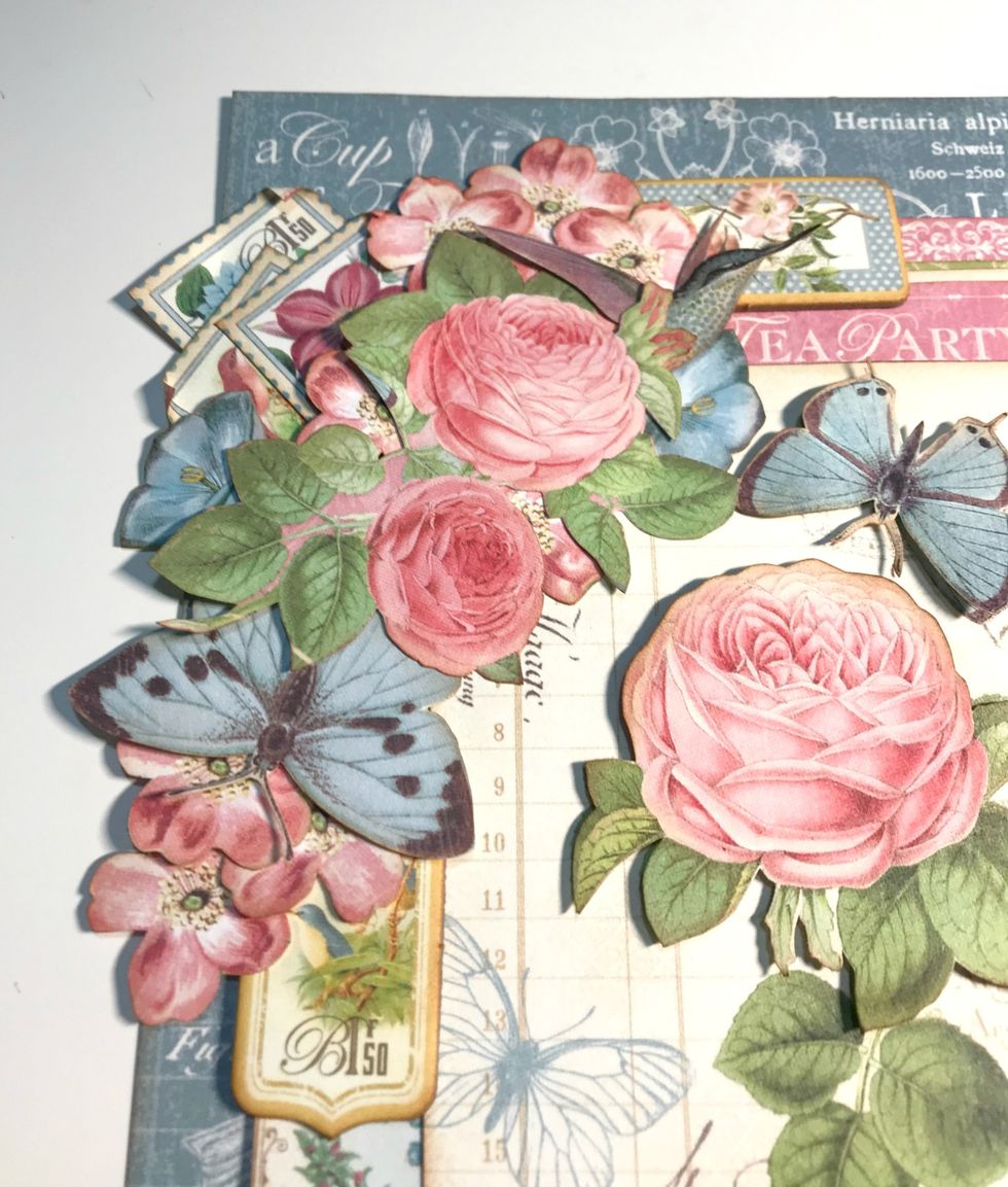 To the right of the large rose, adhere a 1 blue flower & hummingbird. Above the roses, adhere three stamps. Foam square a pink flower between roses & stamps. Roll down a corner on each stamp.