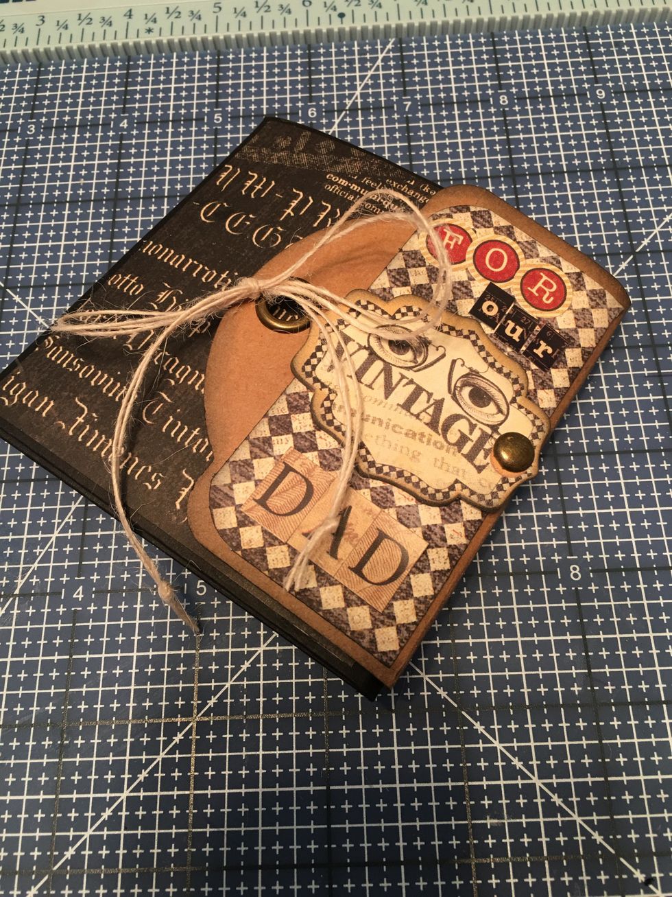 To the "vintage" chipboard piece, insert a brad and cut the long prongs off if needed. Glue the piece to the center of the front cover flap. Add sticker letters for the title "For Our Dad" as shown