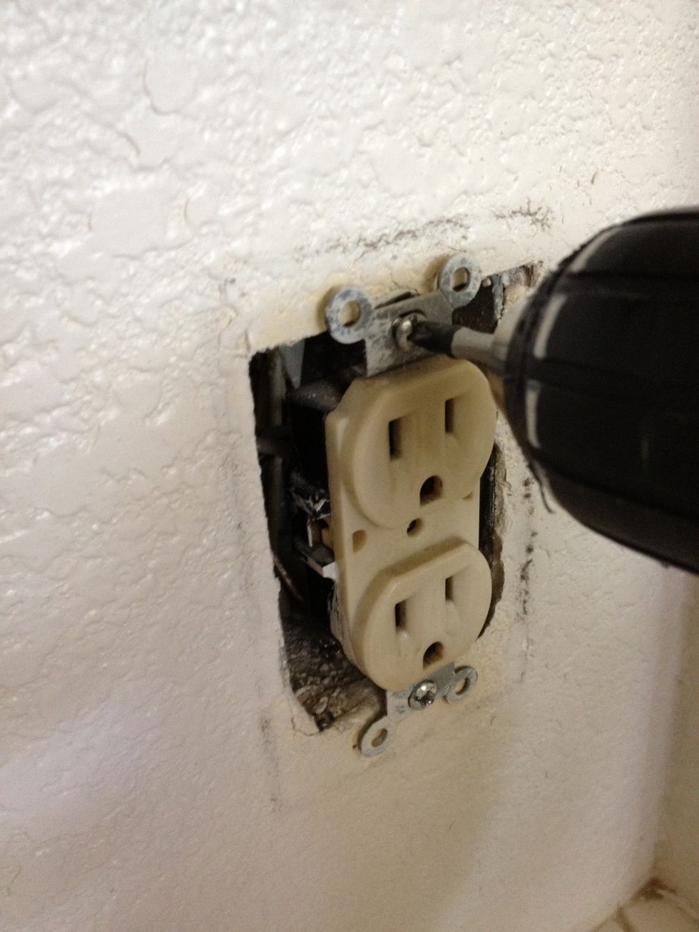 Use a Phillips screwdriver (hand or drill powered based on your personal preference) to remove the screws that hold the outlet to the junction box (the J-box is where all the wires come in).