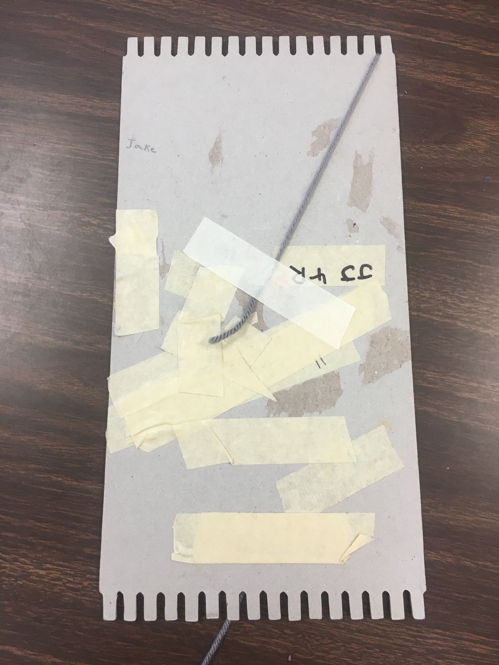Using about a 4 inch piece of masking tape, secure one of the yarn pieces to the back of the cardboard loom. The yarn can still be attached to the roll of yarn at this point.
