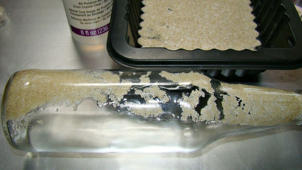 Using an adhesive drip inside the bottle and allow it to run down the side. This will adhere sand to the back side of the bottle behind the rolled message.