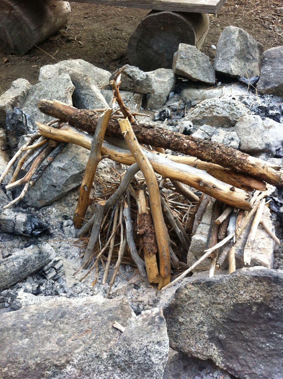 using the stick bridge as support, layer on larger sticks around the pyre. Don't over compact. fire needs breathing room for oxygen. a common error in fire making is smothering it with too much wood.